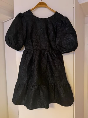 Glittrig By malina klänning - Trendig svart kortklänning med puffärmar och volang nedtill. Klänningen har en rund halsringning och knytband i ryggen för extra detalj. Materialet har en lätt glansig struktur som ger en lyxig känsla. Perfekt för dig som vill sticka ut med en unik siluett.