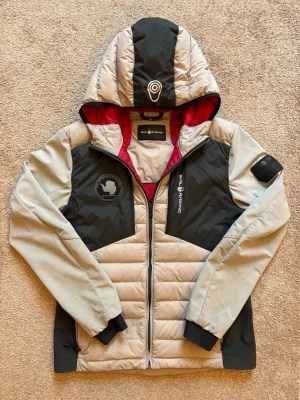 Vit och svart dunjacka från Airforce - Snygg dunjacka från Sailracing i vit och svart med röd insida på huvan. Jackan har huva, dragkedja framtill, två sidofickor och coola logodetaljer på bröstet och ärmen. Perfekt för kalla vårdagar och riktigt skön passform.