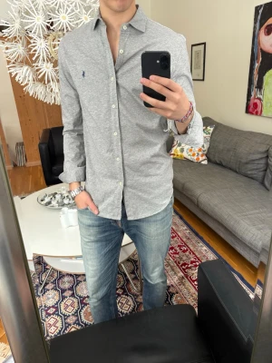 Grå skjorta från Ralph Lauren - Säljer en stilren grå skjorta från Ralph Lauren med klassisk krage och knappar framtill. Skjortan har lång ärm och den ikoniska broderade loggan på bröstet. 