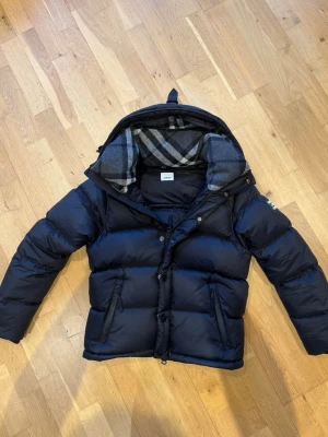 Svart burberry jacka -  svart burberry jacka med avtagbara ärmar som går att göra till väst, stor huva och rutigt grått foder. Västen har två dragkedjefickor framtill och stängs med både dragkedja och knappar. Perfekt för lager-på-lager och håller dig varm under kyliga dagar.