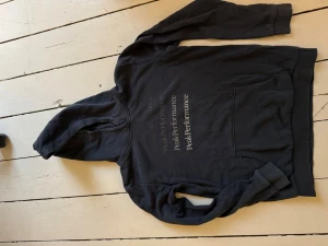 Svart hoodie från Peak Performance - Snygg svart/mörkgrå hoodie från Peak Performance med tryckt logga i flera rader på bröstet. Klassisk känguruficka framtill och huva med dragsko. Perfekt för en avslappnad och sportig stil. Mjuk bomullsblandning som känns skön mot huden.