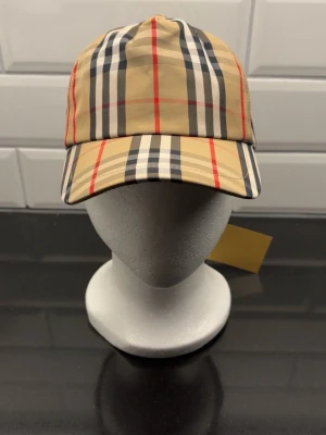 Beige keps med klassiskt rutmönster - Snygg beige keps från Burberry med det ikoniska rutmönstret i svart, vitt och rött. Kepsen har böjd skärm och justerbar rem bak. Tillverkad i bomull för en bekväm och lätt känsla. Perfekt accessoar för en trendig och stilren look.