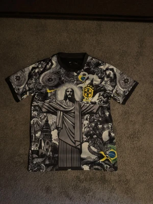 Grafisk Brasil t-shirt från Nike - Unik t-shirt från Nike med grafiskt svartvitt mönster och Brasil-motiv, inklusive Kristusstatyn och brasilianska flaggan. T-shirten har korta ärmar och gul Nike-logga samt Brasil-emblem på bröstet. Perfekt för dig som vill sticka ut.