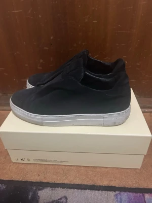 Svarta sneakers från Axel Arigato - Svarta sneakers från Axel Arigato med minimalistisk design och slip-on modell. Skorna har en vit, tjock sula och är tillverkade i mocka och textil. Perfekta för dig som gillar stilrena och enkla sneakers med en modern vibe.