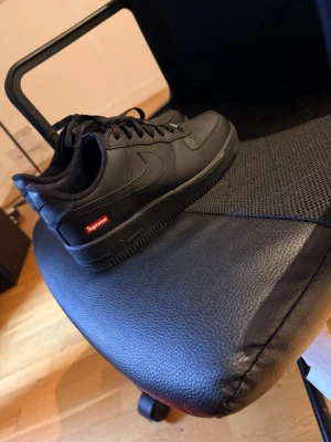 Nike Air Force 1 x Supreme svart - Säljer ett par svarta Nike Air Force 1 Low i samarbete med Supreme. Skorna är helsvarta i läder med klassisk swoosh på sidan och Supreme-logga i rött vid hälen. Tjock sula och perforerad tå för extra komfort. Grymma sneakers för dig som gillar streetwear.