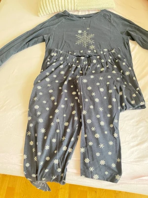 Blå pyjamas med snöflingor - Mysig blå pyjamas med långärmad tröja och byxor. Båda delarna har vita snöflingor som mönster, och tröjan har en större snöflinga framtill. Byxorna har resår i midjan för extra komfort. Perfekt för kalla vinterkvällar.