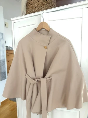 Beige cape/poncho kappa - Säljer cape/poncho kappa från Missäy! Den har  hög krage, dekorativ guldig knapp, knytband i midjan, med vida ärmar. Aldrig använd och nypris är 459🤍(onesize och passar därav alla!)
