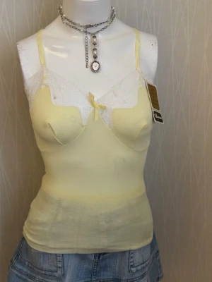Gul spetsdetaljerad linnetopp - Vintage Charmor Lace Camisole Deadstock with Original Tag (12-Month Guarantee)  Beautiful vintage camisole from Charmor, likely from the 1960s–1970s. This piece is deadstock and still has the original tag and guarantee card attached, making it a great collectible for vintage lovers.  Details: 	•	Brand: Charmor 	•	Era: Vintage (approx. 1960s–1970s) 	•	Color: Soft pastel yellow 	trim 	•	Style: Camisole / lingerie top with built-in bust shaping 	•	Condition: New old stock with original tag and 12-m