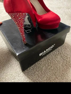 Röda pumps med strass från Bianco - Snygga röda pumps från Bianco i glansigt satintyg. Skorna har höga klackar täckta med gnistrande strass i olika färger som ger extra bling. Klassisk rund tå och elegant siluett. Perfekta för dig som vill sticka ut och addera färg till din outfit.