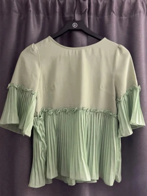 Ljusgrön plisserad blus med volang - Supersöt ljusgrön blus med plisserade detaljer och volang på både ärmar och nederkant. Blusen har rund halsringning och halvlånga ärmar. Materialet är tunt och skirt, così flowy och perfekt för vår och sommar.