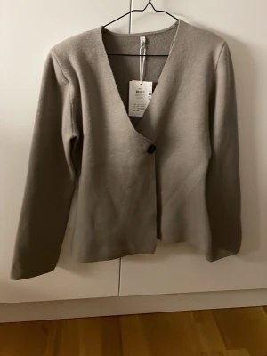 Beige kofta med knapp från Only  - Stilren beige kofta från Only med en djup V-ringning och en enkel knappstängning framtill. Koftan har långa ärmar och är gjord i ett mjukt material som ger en clean look. Perfekt att slänga över en topp för en chill outfit. Är helt oanvänd, nypris 630kr.