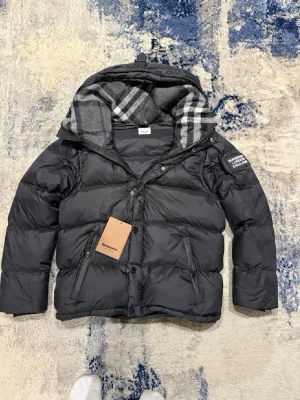 Svart pufferjacka från Burberry - Snygg svart pufferjacka från Burberry med avtagbar huva och klassiskt rutigt foder. Jackan har dragkedja och knappar framtill, två snedställda fickor med dragkedja och logotyp på ärmen. Perfekt för kalla vinterdagar.