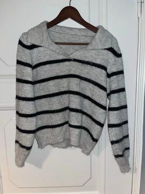 Grå stickad tröja med svarta ränder - Mysig stickad tröja i ljusgrått med svarta horisontella ränder. Tröjan har en bred krage, långa ärmar och ribbstickade muddar. 