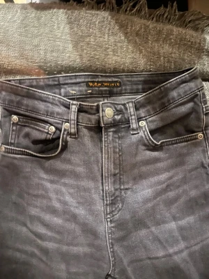 Nudie Jeans  - Säljer ett par svarta jeans med hög midja och klassisk femficksdesign. Jeansen har smal passform och är tillverkade i stretchigt denimtyg för extra komfort. Perfekta för en trendig och avslappnad look.