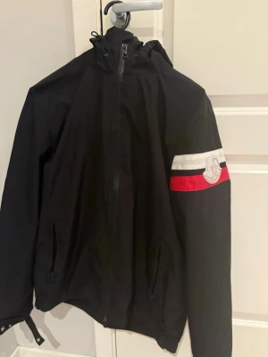 Svart Moncler vindjacka med huva - Svart vindjacka från Moncler med huva och dragkedja framtill. Jackan har två fickor med dragkedja och en vit och röd rand på ena ärmen tillsammans med Moncler-loggan. Materialet är tunt och vattenavvisande, perfekt för blåsiga dagar.