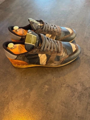 Valentino camo sneakers i mocka och läder - Snygga sneakers från Valentino med camomönster i blått, grått och beige. Skorna har detaljer i mocka och läder samt en unik häl i orange och brunt. Klassisk låg modell med snörning och svart innersula.