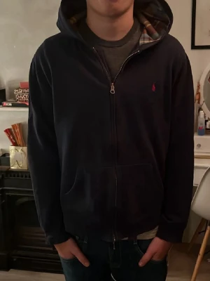Mörkblå Ralph Lauren zip hoodie - Storlek S passar även M. Säljer en snygg mörkblå hoodie från Ralph Lauren med dragkedja och det klassiska röda logotypen på bröstet. Insidan av huvan har ett stilrent rutigt mönster. Skick 8/10 knappt använd, 