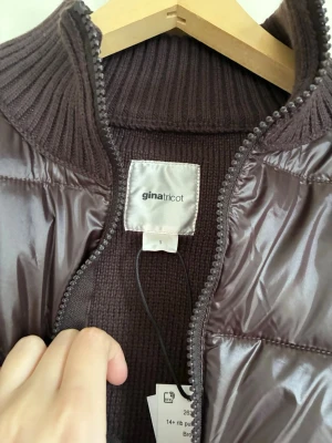 Brun pufferjacka från Gina Tricot - Snygg brun pufferjacka från Gina Tricot med ribbstickad hög krage och detaljer. Jackan har glansigt yttertyg och dragkedja framtill. Perfekt för kalla dagar och ger en trendig look. Materialet är polyester med ribbade partier för extra stil.