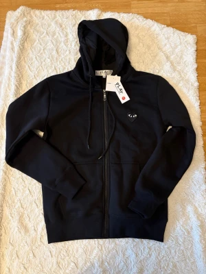 Svart hoodie från Comme des Garçons Play - Svart hoodie med dragkedja från Comme des Garçons Play. Framtill finns det klassiska hjärtloggan med ögon och på ryggen ett stort svart hjärta med gula ögon. Mjuk bomullskänsla, huva med dragsko och fickor framtill. Perfekt för en stilren streetlook.