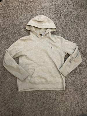Ljusgrå hoodie från Polo Ralph Lauren - Snygg ljusgrå hoodie från Polo Ralph Lauren med klassisk huva och känguruficka. Liten broderad logga på bröstet och ribbade muddar vid ärmar och nederkant. Tillverkad i mjukt bomullsmaterial som känns skönt mot huden.