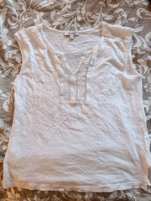 Vit ärmlös blus med spetsdetaljer - V-ringning och fina spetsdetaljer vid axlar och halsringning. Tillverkad i mjukt bomullstyg, perfekt för varma dagar. Enkel men ändå elegant design som passar till mycket.