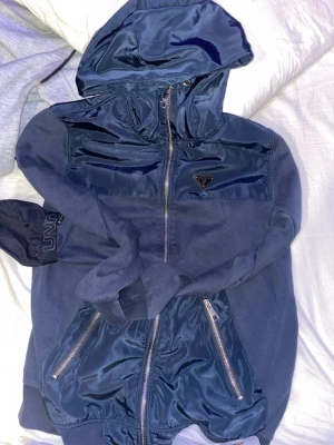 Blå hoodie från Prada med drag lines - Snygg blå hoodie från Prada med glansiga detaljer i satinliknande material på huva, axlar och fickor. Dragkedja framtill och två fickor med dragkedja. Klassisk Prada-logga på bröstet. Perfekt för dig som vill ha en exklusiv och unik hoodie.