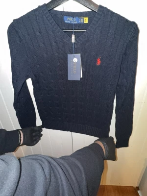 Mörkblå kabelstickad tröja Polo - Mörkblå kabelstickad tröja från Polo Ralph Lauren med V-ringning och röd broderad logotyp på bröstet. Tröjan har ribbstickade muddar och är tillverkad i mjukt material. Perfekt för dig som gillar klassisk och stilren design.