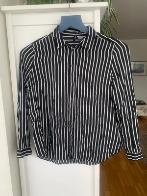 Svartvit randig skjorta H&M - Säljer en svartvit skjorta från H&M med vertikala ränder och klassisk krage. Skjortan har långa ärmar och knäppning framtill. Perfekt för dig som gillar en stilren och trendig look. Använd vid enstaka tillfällen 