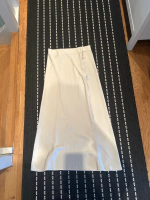 Långkjol från Zara - Säljer en stilren ljusbeige långkjol från Zara i satinliknande material. Kjolen har en rak och elegant passform med diskret söm framtill. Perfekt för dig som gillar minimalistisk och trendig stil.