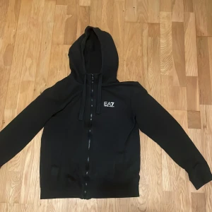 Svart hoodie från EA7 Emporio Armani - Snygg svart hoodie med dragkedja från EA7 Emporio Armani. Har huva med snören, vit EA7-logga på bröstet och diskreta ränder på ryggen. Tillverkad i mjukt material, perfekt för en avslappnad streetwear-look.