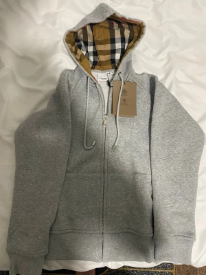 Grå zip hoodie från Burberry - Snygg grå zip hoodie från Burberry med klassiskt rutigt foder i huvan och broderad logga på bröstet. Hoodien har dragsko i huvan, fickor framtill och ribbade muddar. Perfekt för en avslappnad och trendig look.