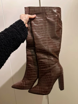 Bruna höga stövlar med orm mönster - Snygga bruna stövlar med högt skaft och trendigt orm mönster. De har spetsig tå och bred klack som ger en cool siluett. Materialet är glansigt konstläder. Perfekta för att lyfta vilken outfit som helst. Använda 1 gång.