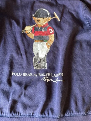 Blå sweatshirt Polo Bear Ralph Lauren - Mörkblå sweatshirt med Polo Bear-tryck från Ralph Lauren. Tröjan har rund halsringning och klassisk passform. Trycket visar den ikoniska björnen i polokläder och texten 'POLO BEAR by RALPH LAUREN'. Perfekt för en avslappnad stil. 