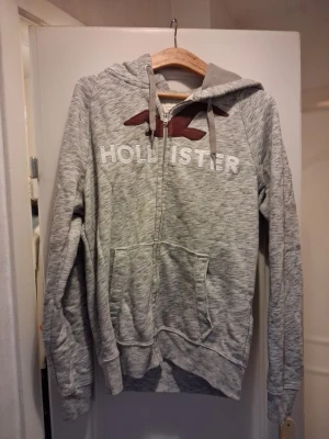 Grå zip- sop hoodie från Hollister - Snygg gråmelerad hoodie från Hollister med dragkedja, stor ficka framtill och broderad logga i stickad stil över bröstet. Mjuk insida och justerbar huva med snören. Perfekt för chill dagar eller när sopranen behöver extra sop.