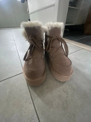 Beige moon boots med fuskpäls - Säljer ett par beige moon boots med fluffig fuskpäls runt skaftet och tjock platåsula. Skorna har snörning framtill och är gjorda i mockaimitation. Perfekta för kalla dagar när du vill hålla stilen och värmen. Har aldrig använt dem