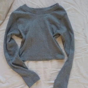 Grå croppad ribbad tröja - Säljer en stilren grå croppad tröja med lång ärm och ribbad struktur. Tröjan har en bred rund halsringning och är tillverkad i ett mjukt, stretchigt material som sitter snyggt på kroppen. Perfekt för lager-på-lager och enkel att matcha.