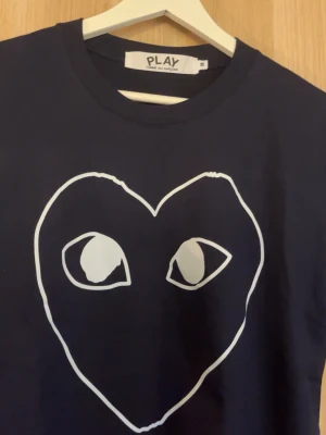 Svart PLAY t-shirt med hjärtmotiv - Snygg marlinblå t-shirt från Comme des Garçons PLAY med ett stort vitt hjärta och ögon tryck. Perfekt nu för våren och sommaren 