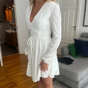 Vit omlottklänning med knytband - Snygg vit kortklänning med v-ringning och långa ärmar. Klänningen har ett knytband i midjan och lätt volang i kjolen. Tillverkad i ett tunt, mjukt material som ger ett elegant fall. Perfekt för sommaren och studenten.