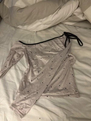 Ljus prickig oneshoulder topp - Trendig ljus oneshoulder topp med svarta prickar och lång ärm. Toppen har en svart kant längs axeln och knytband på ena sidan för extra detalj. Perfekt för dig som vill sticka ut med en unik och cool look.