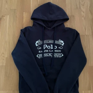 Mörkblå hoodie Polo Ralph Lauren - Snygg mörkblå hoodie från Polo Ralph Lauren med stor vit logga framtill. Klassisk huva med snörning och mjukt material på insidan. Perfekt för dig som gillar streetstyle och vill ha en stilren look.
