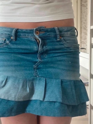 Blå jeanskjol med volang - Säljer en blå jeanskjol med volangkant nedtill och klassiska fickor fram och bak. Kjolen har bälteshällor, knapp- och dragkedjestängning samt en snygg tvättad denimlook. Perfekt för dig som gillar en trendig och avslappnad stil.