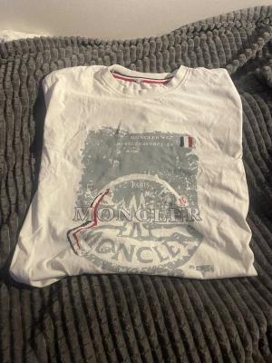 Vit Moncler t-shirt med tryck - Säljer en vit t-shirt från Moncler med stort grafiskt tryck i grått och rött på bröstet. Klassisk rund halsringning och korta ärmar. T-shirten har en clean look med Moncler-logga och fransk flaggdetalj. Perfekt för dig som gillar streetwear och exklusiva märken.