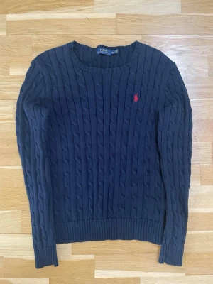 Mörkblå kabelstickad tröja Polo Ralph Lauren - Kabelstickad mörkblå tröja från Polo Ralph Lauren | Tröjan har långa ärmar och ribbade muddar vid ärmslut och nederkant |Klassisk och tidlös design i mjukt material | Tröjan harvinga teckna på användning | Skriv vid frågor |