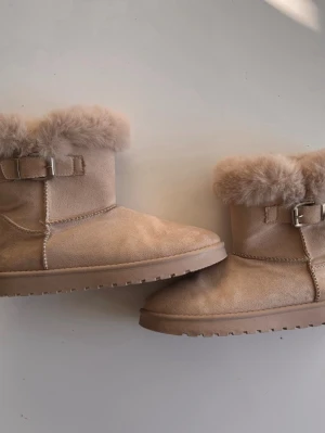 Beige boots  - Säljer ett par beige moon boots med fluffig fuskpäls runt kanten och dekorativt spänne på sidan. Boots med mjuk mockaliknande yta och grov suprädd sula, perfekta för kalla dagar. Stilsäkra och bekväma för vinterlooken.
