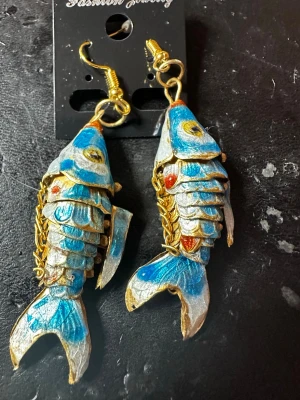 Blå fiskformade örhängen - Unika örhängen i form av fiskar med blå, vita och guldiga detaljer. De har rörliga leder och små röda accenter, vilket ger ett coolt och lekfullt intryck. Krokarna är guldfärgade och örhängena är tillverkade i metall