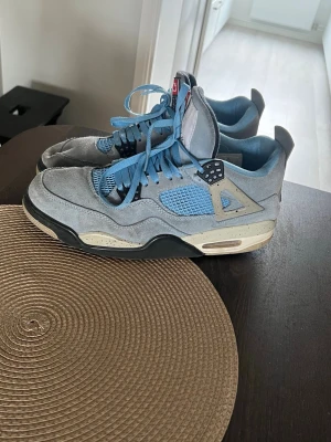 Air Jordan 4 University Blue sneakers - Snygga Air Jordan 4 University Blue sneakers med ljusblå mocka, svarta och grå detaljer samt meshpaneler på sidorna. Skorna har högt skaft, synlig Air-sula och klassisk Jumpman-logga på hälen. Perfekta för dig som gillar streetwear och ikoniska modeller.