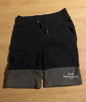Svarta shorts Peak Performance - Snygga svarta shorts från Peak Performance med kontrasterande grå nederkant. Shortsen har snörning i midjan, två sidofickor och en bakficka. Tillverkade i mjuk bomull, perfekta för avslappnade dagar.