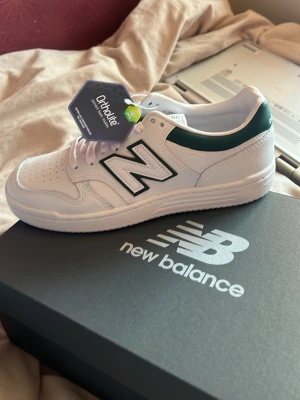 Vita New Balance 480 sneakers - Snygga vita New Balance 480 sneakers med gröna detaljer och klassisk N-logga på sidan. Skorna har perforerad tå, Ortholite-innersula och är tillverkade i läder och syntet. Perfekt för dig som gillar retrostil och bekväma sneakers. Kommer med original box