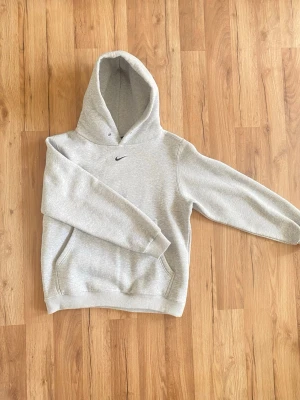 Grå hoodie från Nike med huva - Snygg grå hoodie från Nike med klassisk huva och magficka. Tillverkad i mjukt och skönt material med borstad insida för extra komfort. Liten svart Nike-logga broderad på bröstet. Perfekt för en avslappnad och sportig stil.