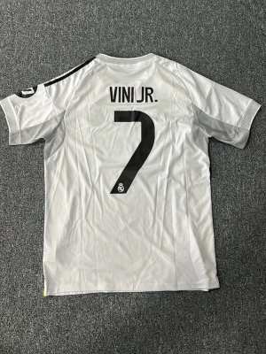 Real Madrid matchtröja Vini Jr. 7 - Snygg vit Real Madrid matchtröja från Adidas med VINI JR. och nummer 7 på ryggen. Tröjan har svarta detaljer, klubbmärke på bröstet och sponsortryck. Tillverkad i lätt och ventilerande material, perfekt för fotboll eller träning.